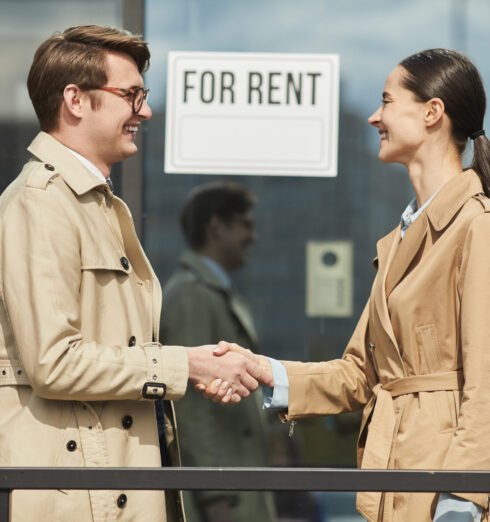 Rental Agent Shaking Hands with Client Side view waist up portrait of smiling real estate agent shaking hands with client outdoors while standing next to FOR RENT sign