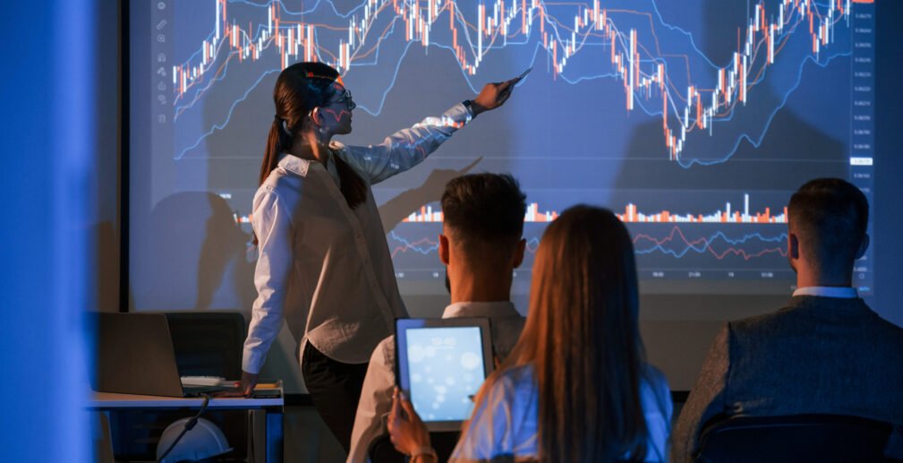 Showing the plan on the projector. Female leader talking to employees in office of stock exchange company Showing the plan on the projector. Female leader talking to employees in office of stock exchange company.