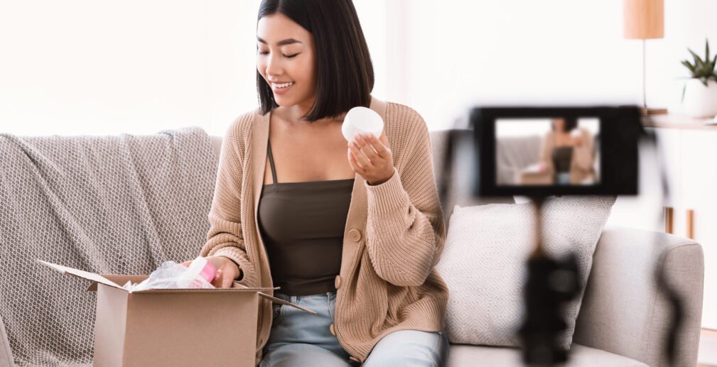 Happy asian woman recording product review for beauty blog Product Review. Satisfied asian young woman vlogger holding moisturizer cream, showing cosmetics products, recording video for her social media blog on camera at home, unpacking cardboard beauty box